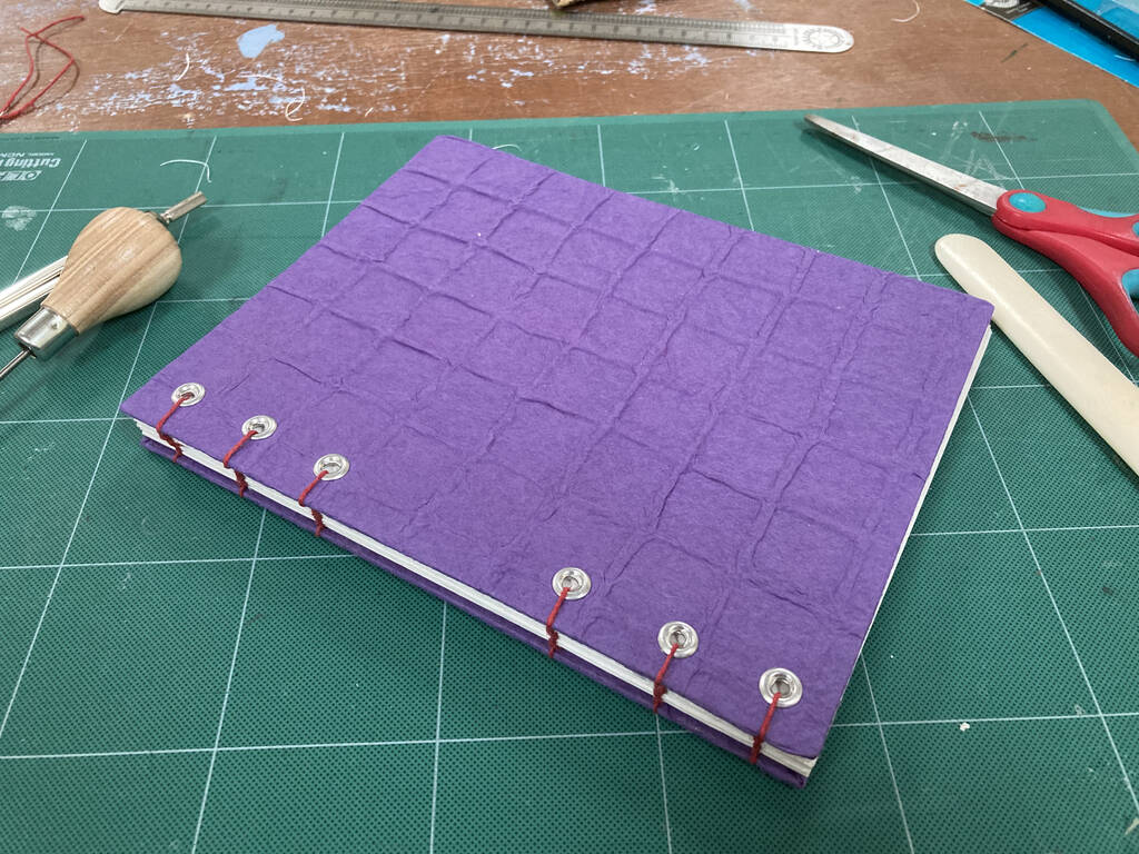 Photo of a coptic-stitched book with a textured purple cover and red stitching, sitting on a green cutting mat.