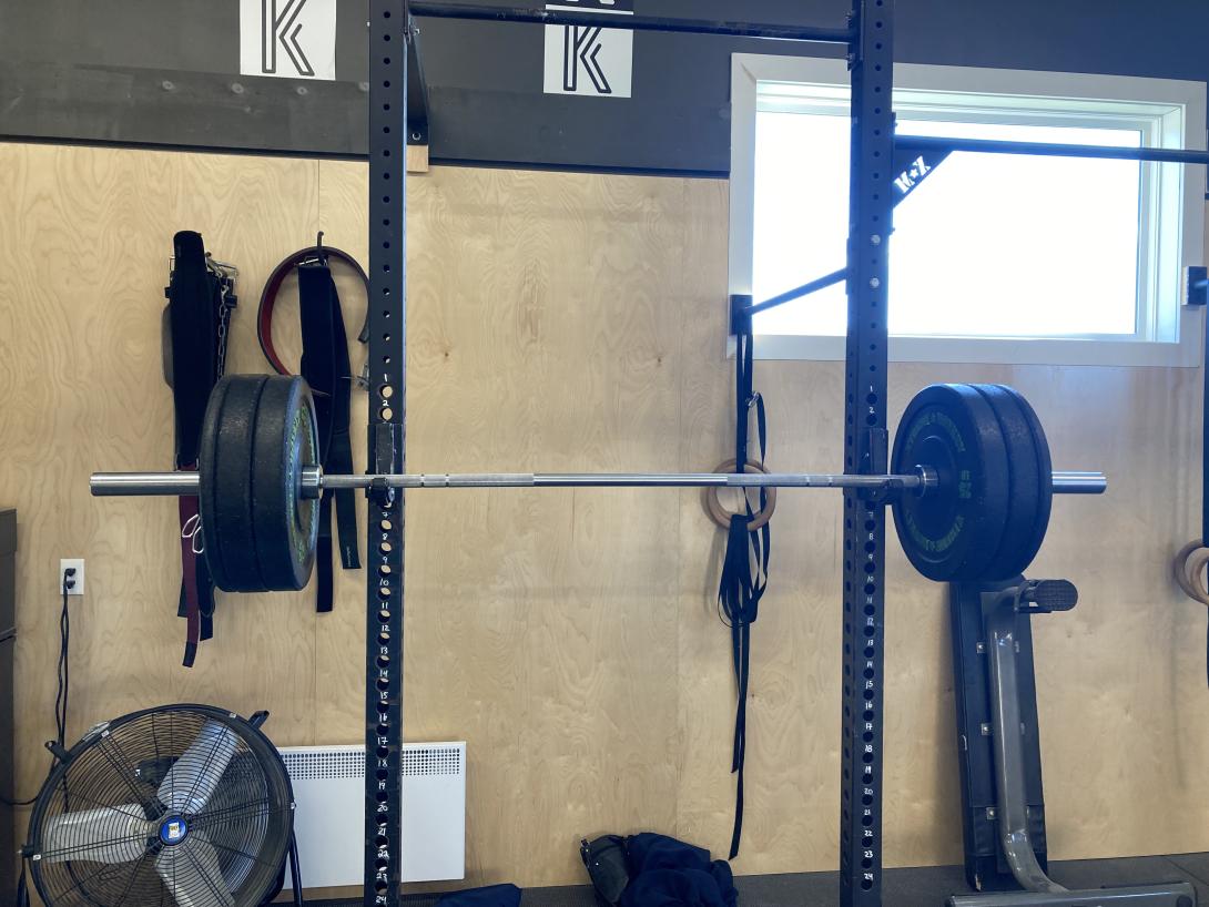 A barbell on a rack, with three weights on each end, in front of a plywood wall, with stylized letter K's above, and a brightly lit window to the top-right.