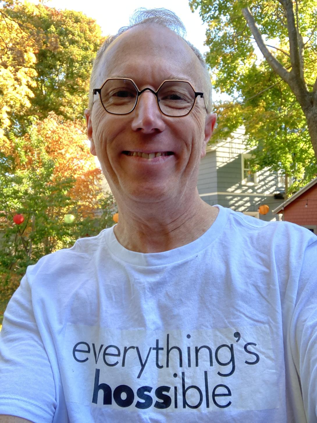 A selfie of Peter Rukavina, taken in front of fall leaves in his back yard, Peter is white man, with short close-cropped hair, wearing orange honeycomb-shaped eyeglasses. He is smiling, and wearing a white T-shirt with the phrase "everything's hossible," all lower case, printed on it, with the "hoss" emphasized.