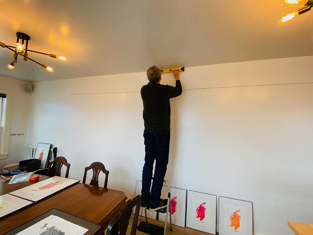 A man wearing dark trousers and a dark sweater standing on a stepladder, arms extended in front of him, holding a yellow level against the corner of the ceiling and wall. On the ground at his feet are a multi-coloured collection of framed prints set against the wall.