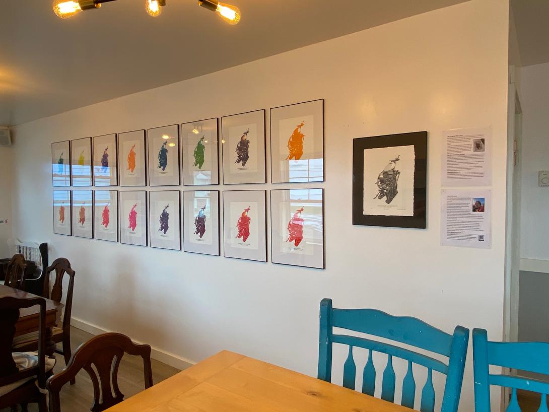 Two rows of framed multicoloured prints hung on a white wall. To the right is a single black and white print, on a black background, and to the right of that are a printed artist statement and description of the work.