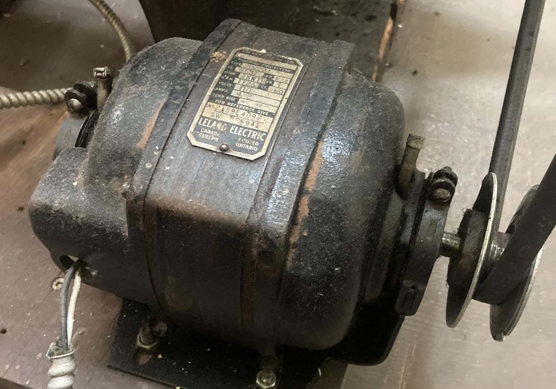 Photo of a small electric motor. There is a nameplate on the top of the motor, with technical details, and LELAND ELECTRIC, Guelph, Canada. On the right side the motor's drive shaft is connected to a drive belt.