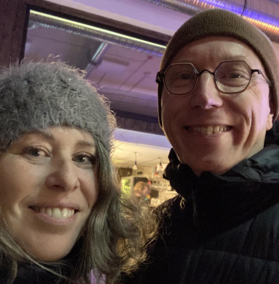 Lisa and I, side by side, in a selfie taken during our trip to Europe. We are both wearing winter coats and winter hats, and we're standing outside, in the evening, in front of a storefront.