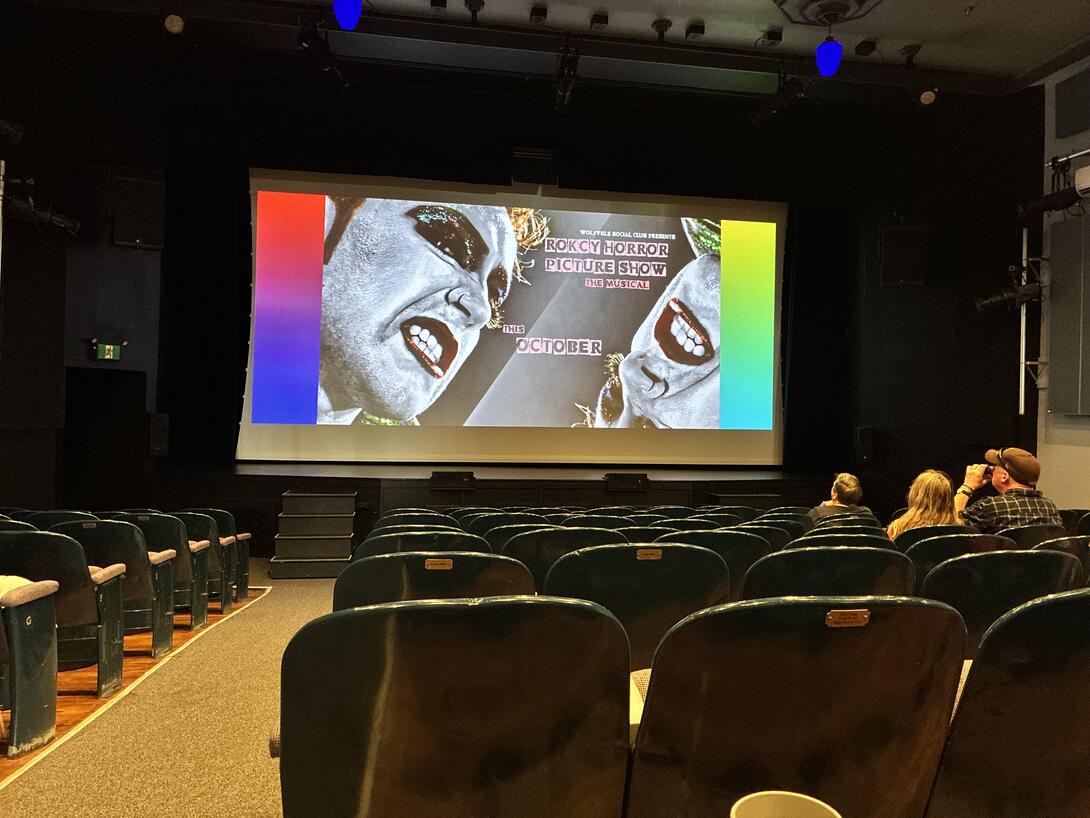A view of an ad for Rocky Horror on the screen of the theatre, with a view of the seats.