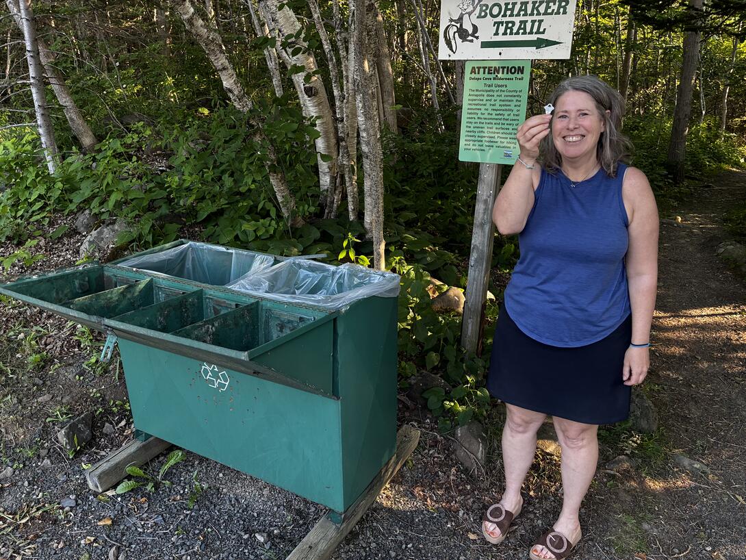Lisa standing to the right of a green garbage bin, open to reveal the bags inside. She's holding a key in her right hand. She is smiling.