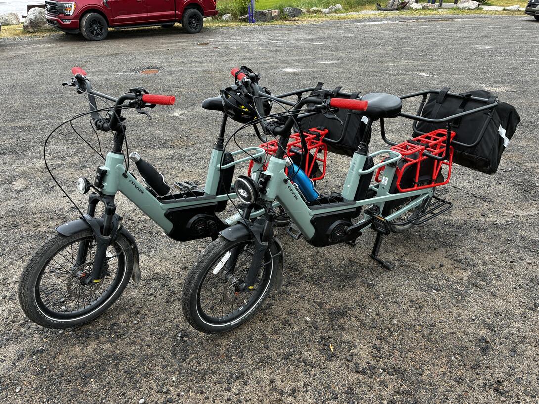 Two Canondale Wargonwagon e-bikes, green with red racks on the back, and rear panniers, side by side, parked.