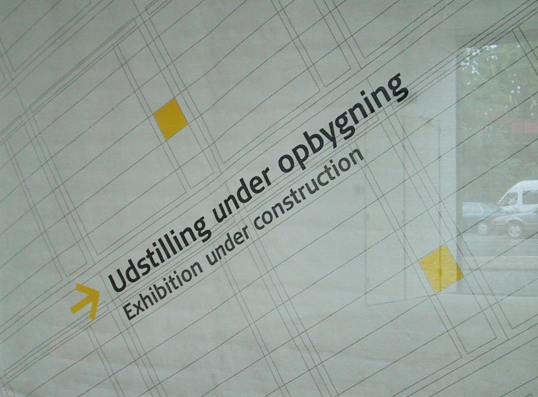 A poster in a window with the text "Exhibition under construction" in English under the Danish equivalent. The poster has spreadsheet-like rows and columns, punctuated by bright yellow squares.