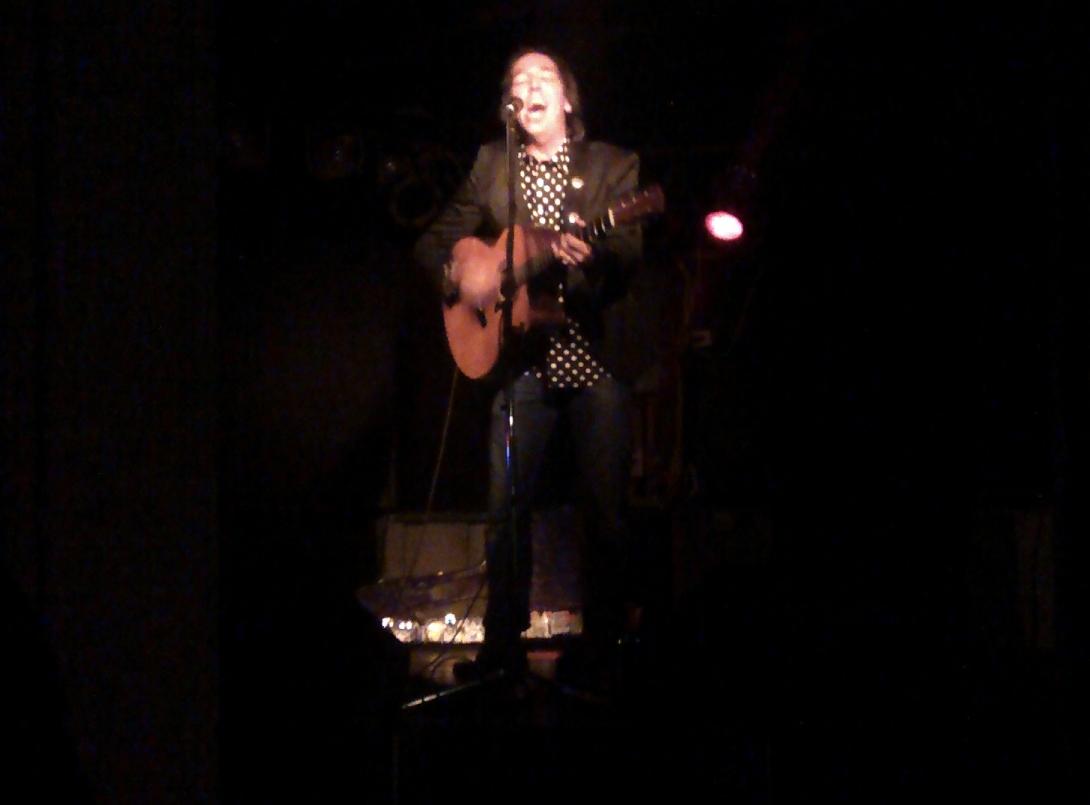 Stephen Fearing, singing on stage in Sackville, NB, in October 2013. He is on stage alone, with a black background, wearing a black and white polkadot shirt, a black jacket, and holding a guitar.