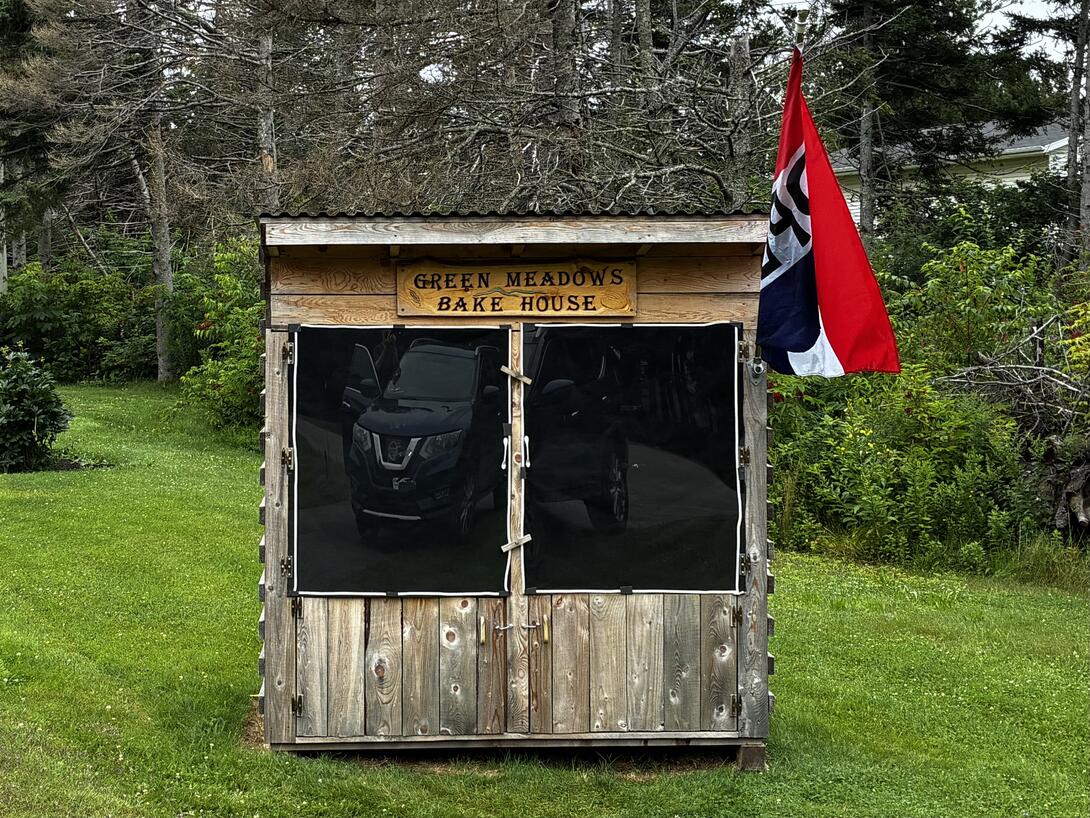 A small wooden shed with large smoky glass doors on the front, with an engraved sign "Green Meadows Bake House" on top.
