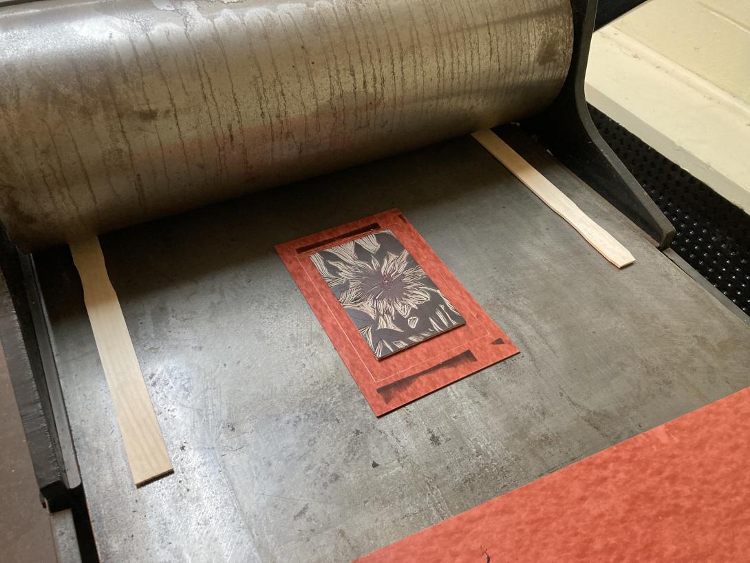 An etching press with a lino block set on red shirt cardboard on the bed, with to paint stir sticks set on either side.