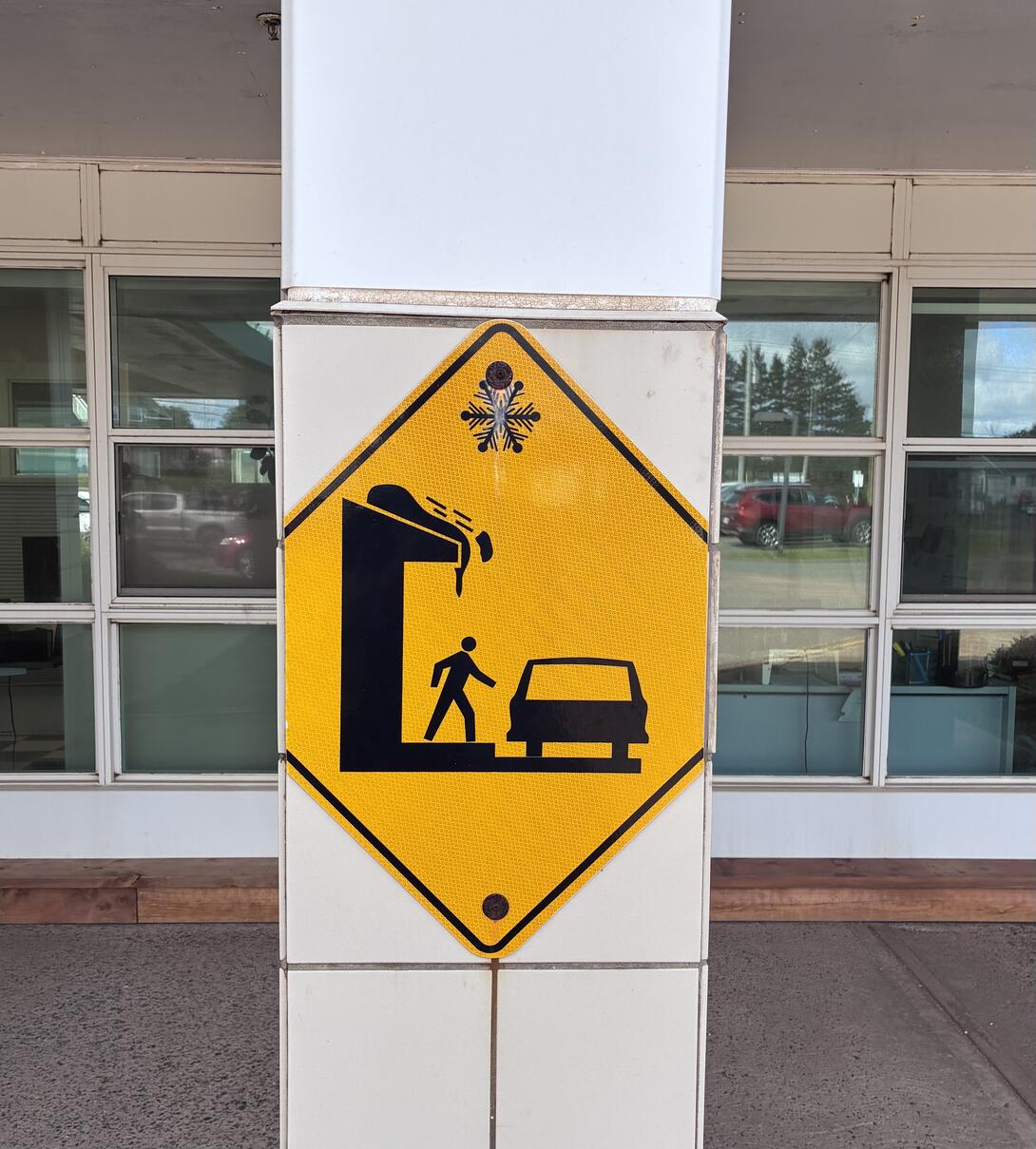 A yellow sign showing a simple graphic of snow falling off a roof and onto the hood of someone getting into a car.
