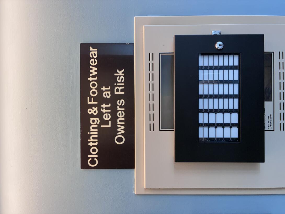 A black sign with "Clothing & Footwear Left at Owners Risk" engraved, above an alarm control panel.