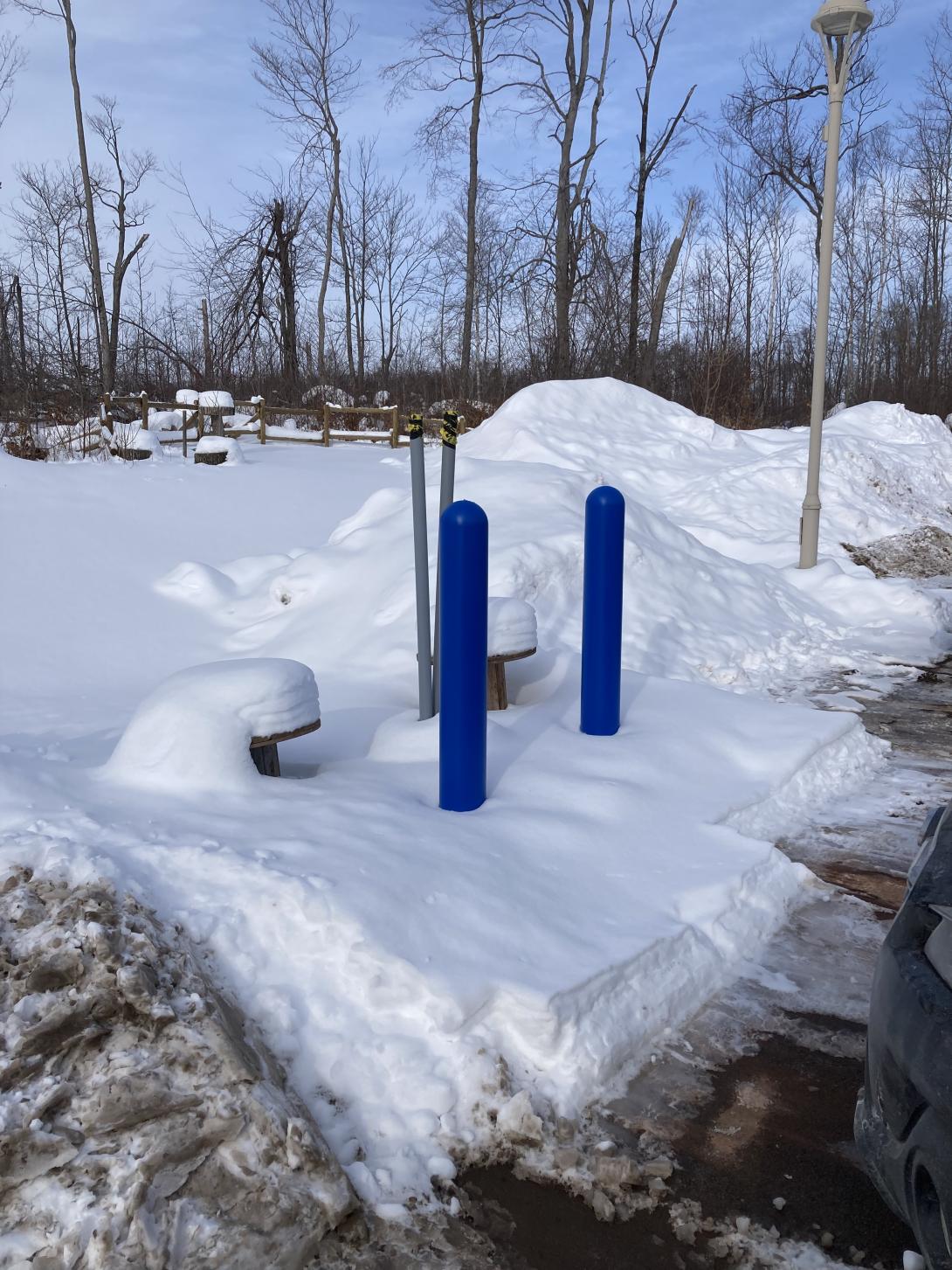 Two grey electrical conduits sticking out of the snowy edge of a parking lot, with two blue bollards in front.