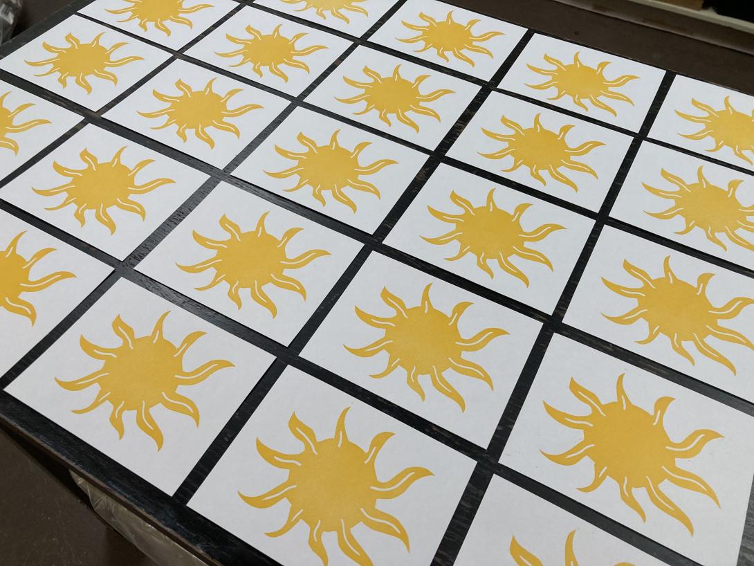 A grid of 25 small cards, each printed with an abstract yellow sun, laid out on a black table to dry.