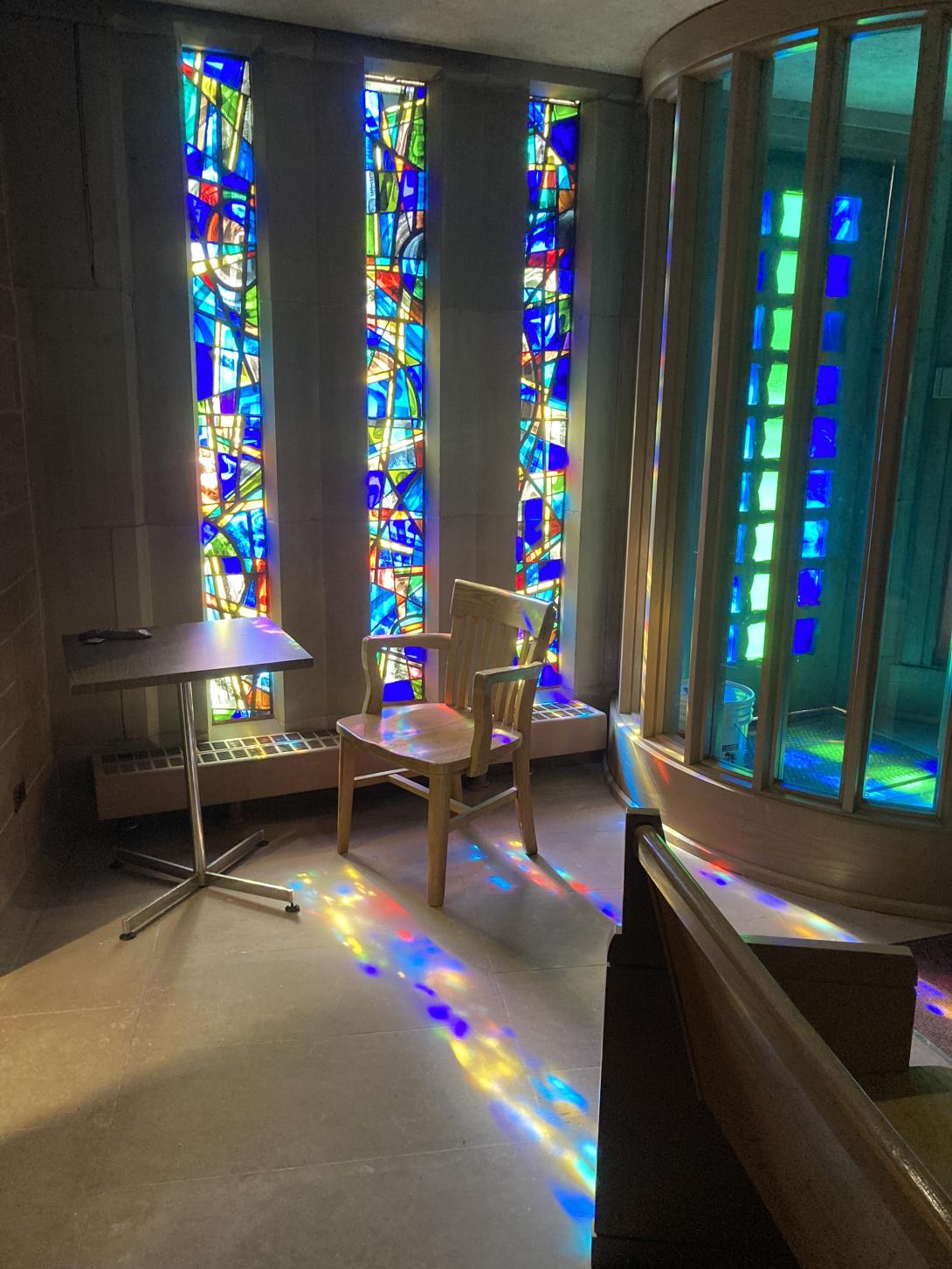 A table and chair in front of vertical stained glass windows, in blue, red, and yellow in abstract patterns. There is light streaming through the windows and patterns of colour on the floor as a result.