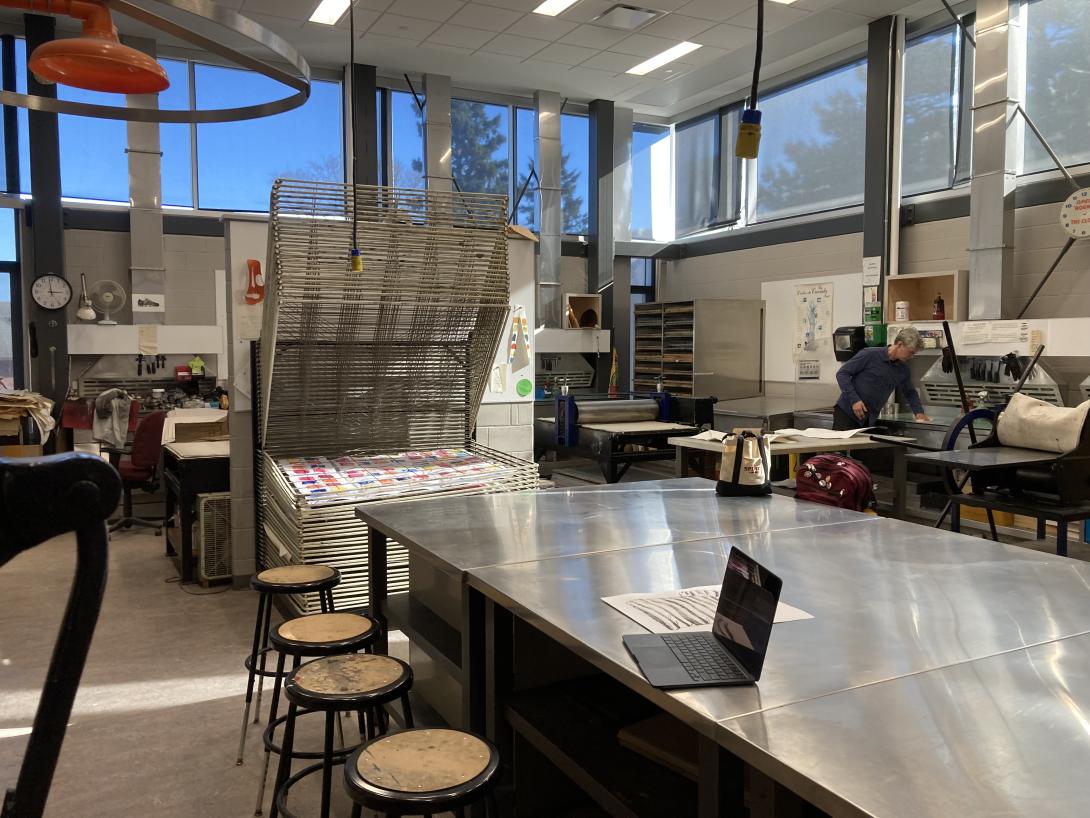 A view of the printmaking shop, with a large metal table in the foreground, a drying rack and presses in the background. The space is filled with light, and there are large windows surrounding.