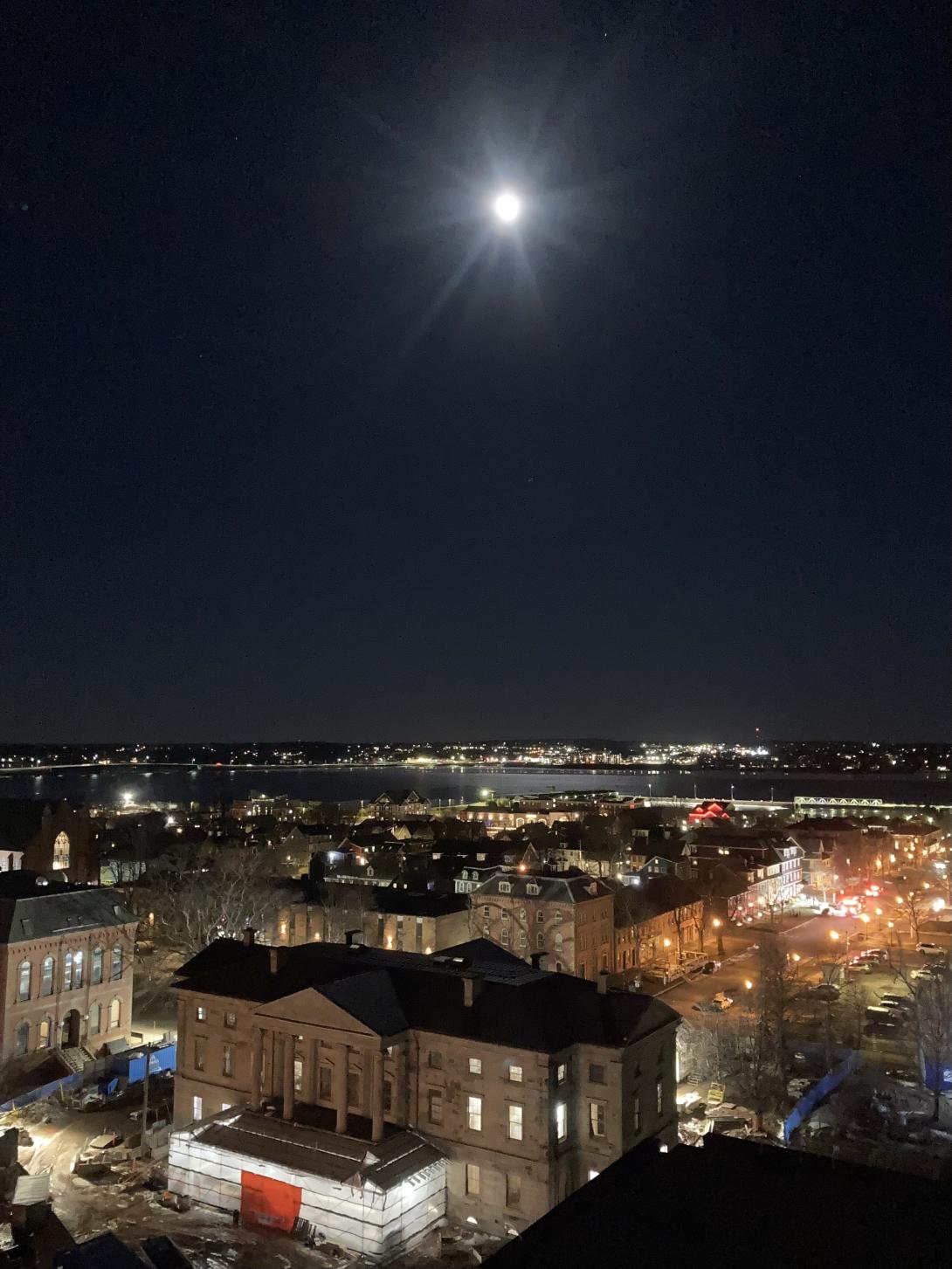 A bright full Moon shining over an illuminated Province House, taken from above, with the lights of downtown in the foreground and Stratford in the background. 