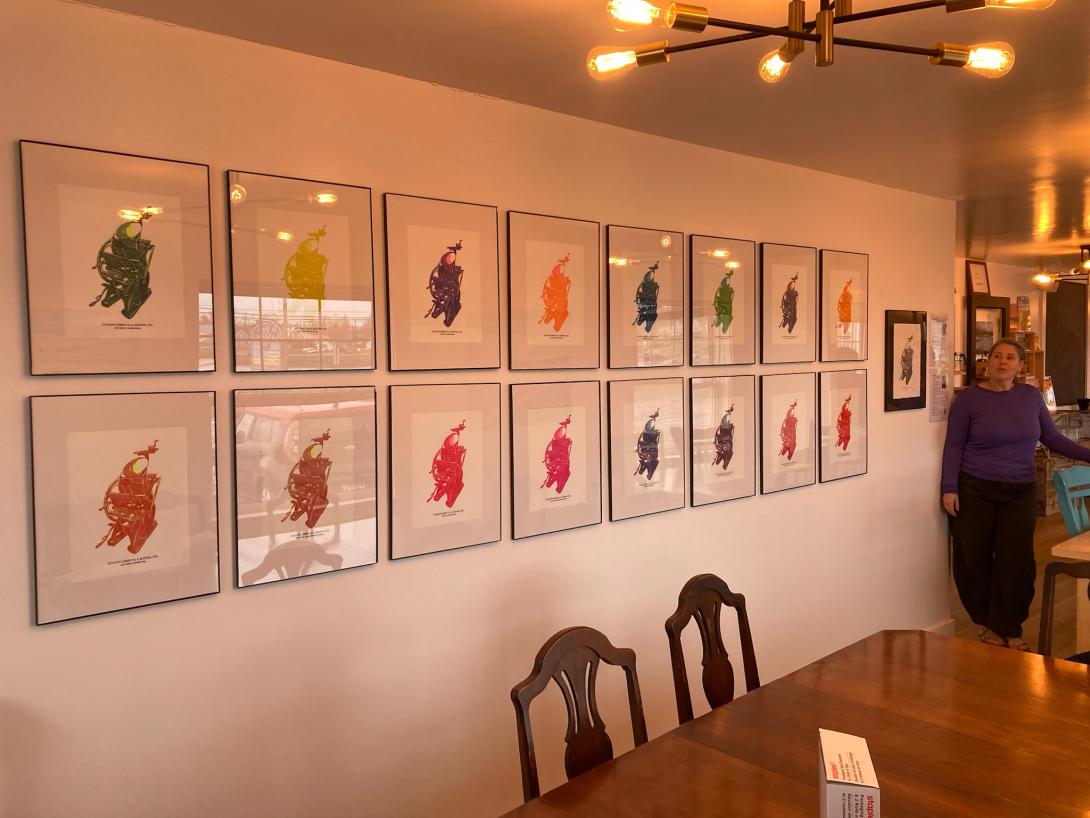 Framed colourful prints hung on a white wall in two rows. A single black and white print sits at the end, and beside it is a woman with long hair tied back, wearing a purple sweater.