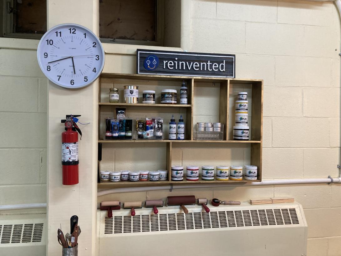 A gold shelf on a cinder block wall, filled with ink bottles. To the left is a clock, showing 5:42 p.m., and a red fire extinguisher. Below is a radiator cover, with ink brayers set on it.