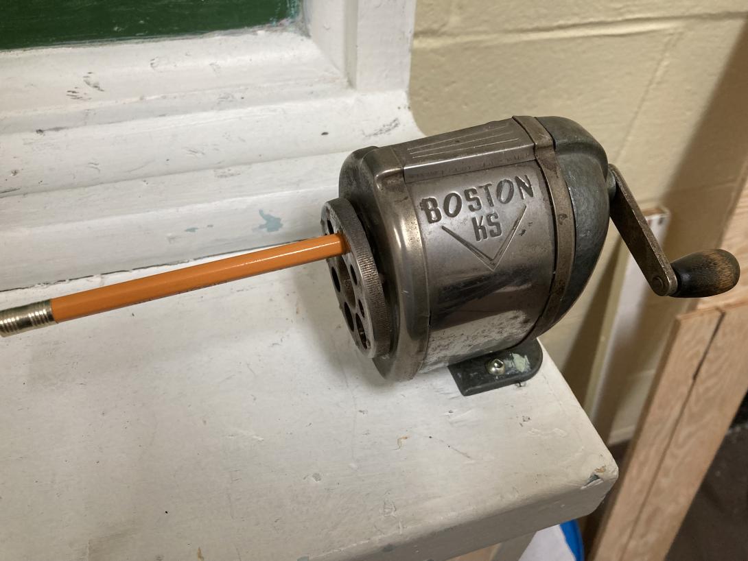 An older rotary pencil sharpener screwed to a white ledge.