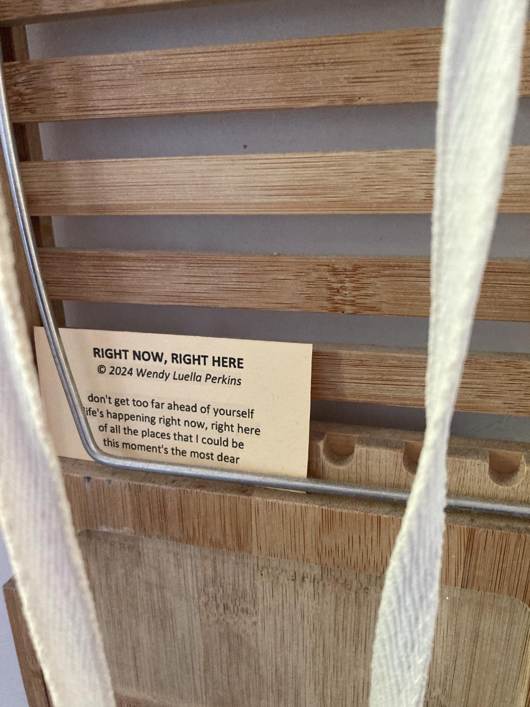 A small card set into a corner of a wooden rack, reading "don't get too far ahead of yourself life's happening right now, right here of all the places that I could be this moment's the most dear"