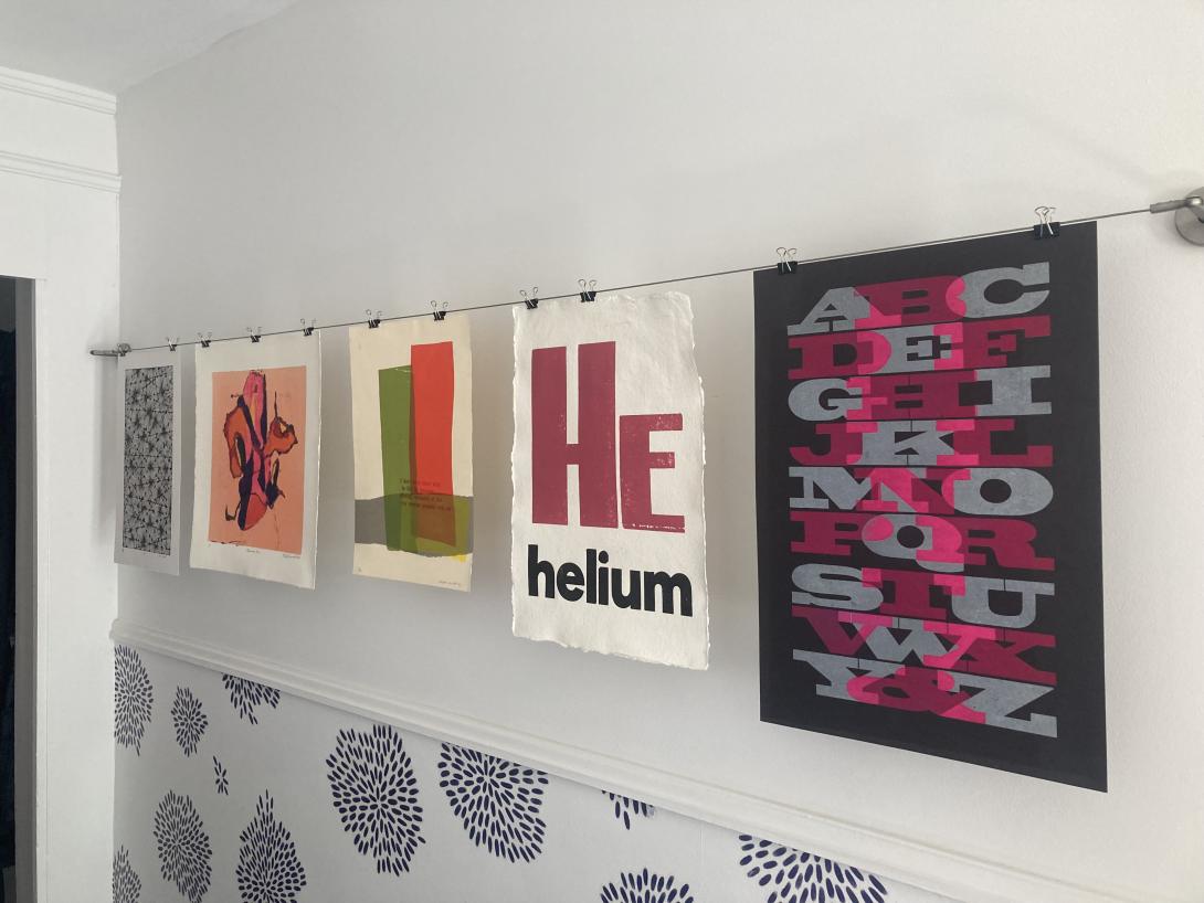 A white hallway with a wire strung across the wall at eye level. There are 5 pieces of art hung on the wire, front left to right: a black and white abstract drawing, a colourful monotype print, a print with three simple coloured bars overlaid with red type, a broadside with bold aubergine HE, with helium printed underneath in black, and a black poster printed with an overlapping alphabet in grey and pink.