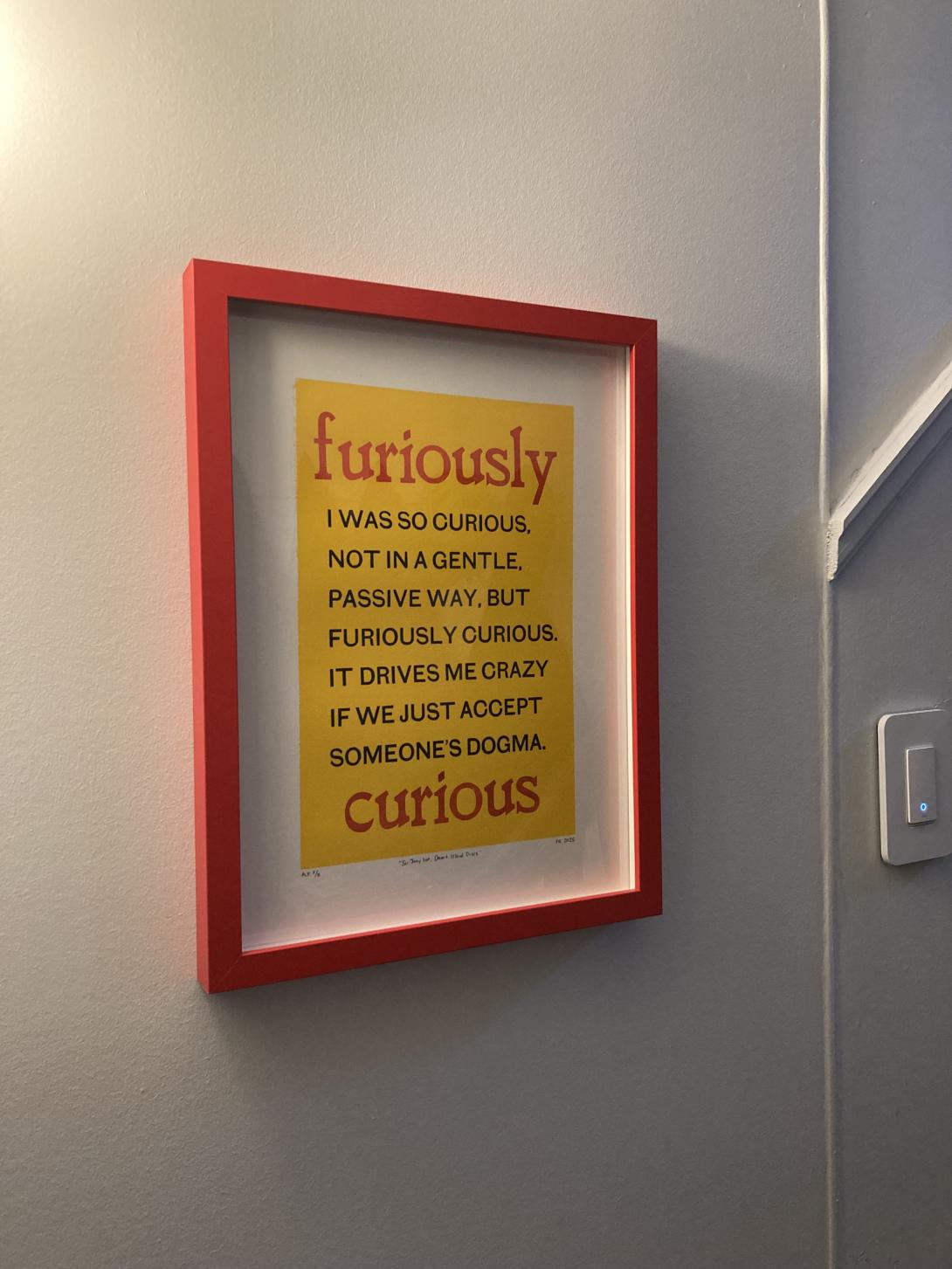 On a white wall, at the bottom of the stairs, lit from the left, is a red-framed broadcast with the words "furiously" and "curious" printed, in lower case, in red, at the top and bottom, with the text "I WAS SO CURIOUS, NOT IN A GENTLE, PASSIVE WAY, BUT FURIOUSLY CURIOUS. IT DRIVES ME CRAZY IF WE JUST ACCEPT SOMEONE'S DOGMA." printed, all caps, in black, between, all on a bright yellow background.