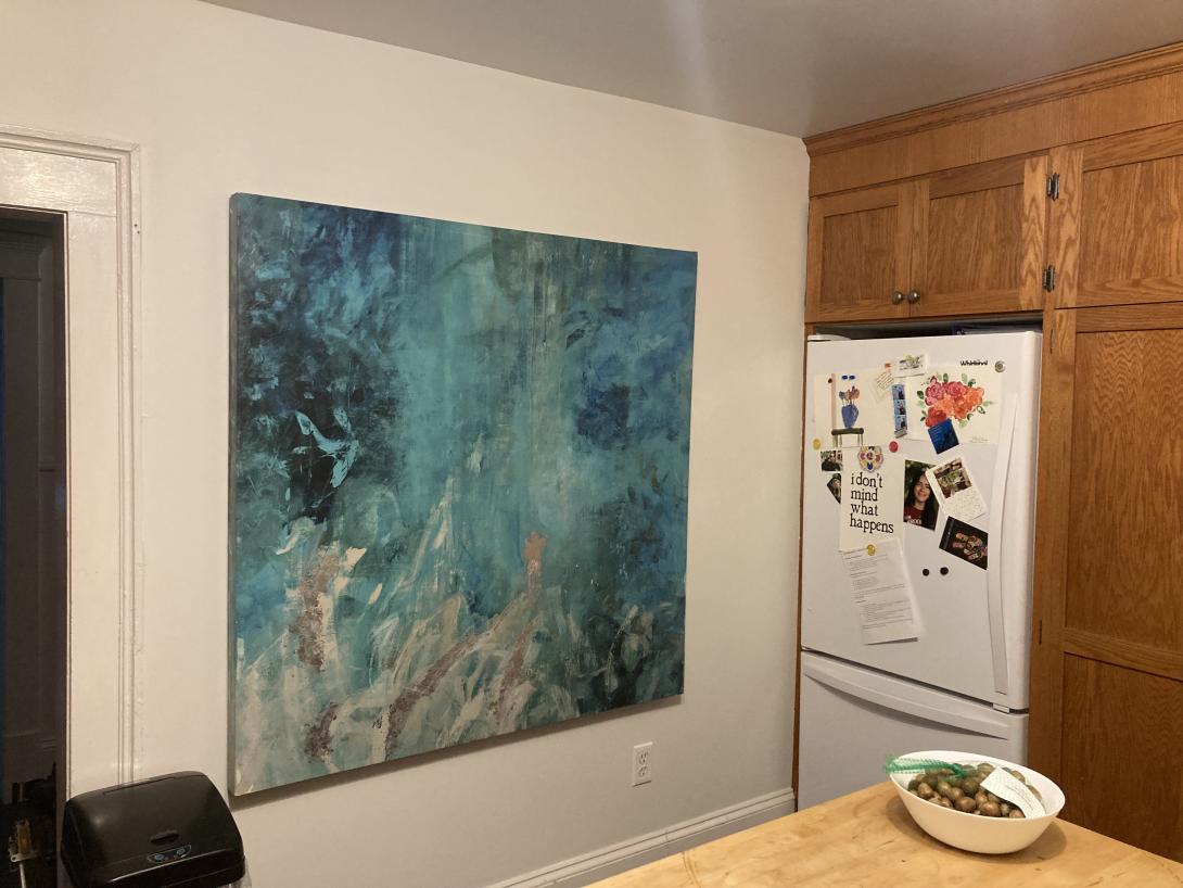 A large abstract painting, in blues, greens, and greys, hung on the white wall of a kitchen, with the fridge, covered in ephemera, to the right.