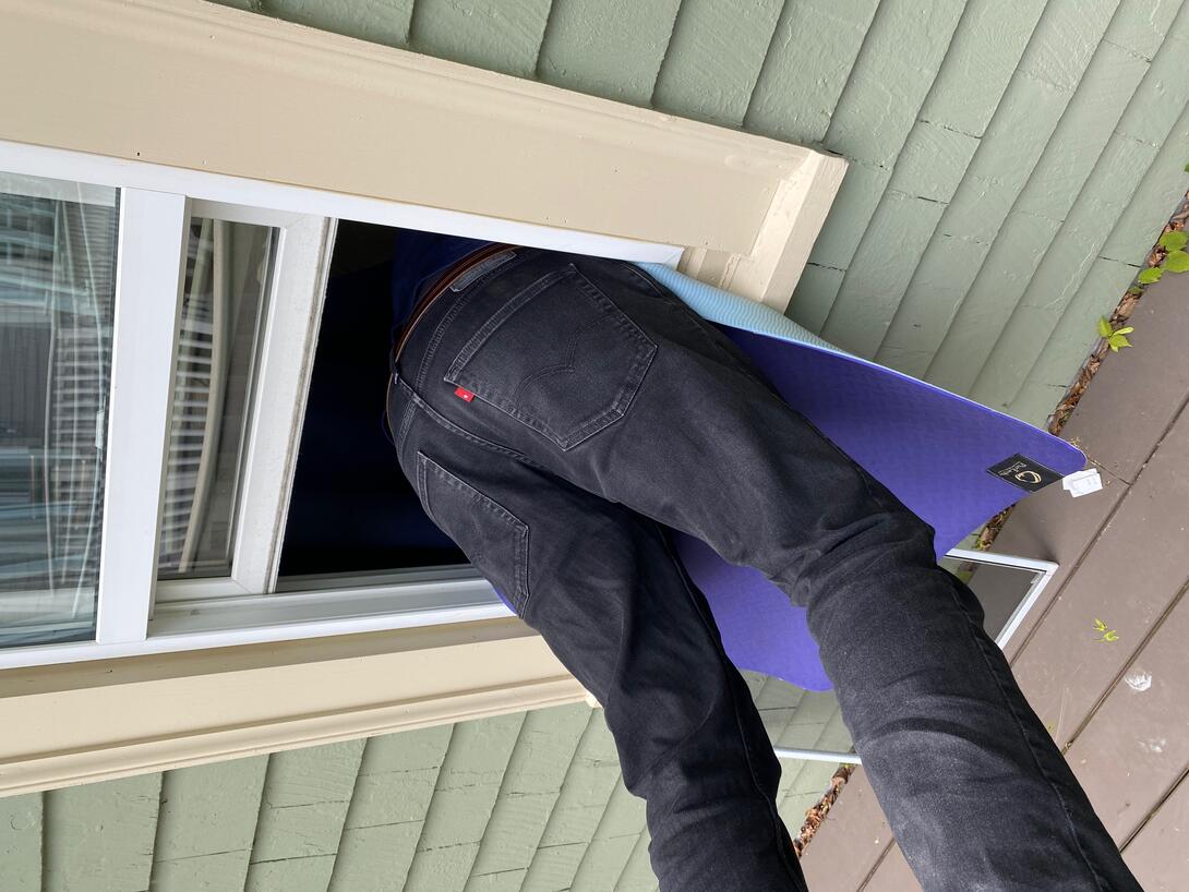 A photo of a man wearing black jeans halfway through an open window. Only his legs are showing. There's a yoga mat protecting his chest.