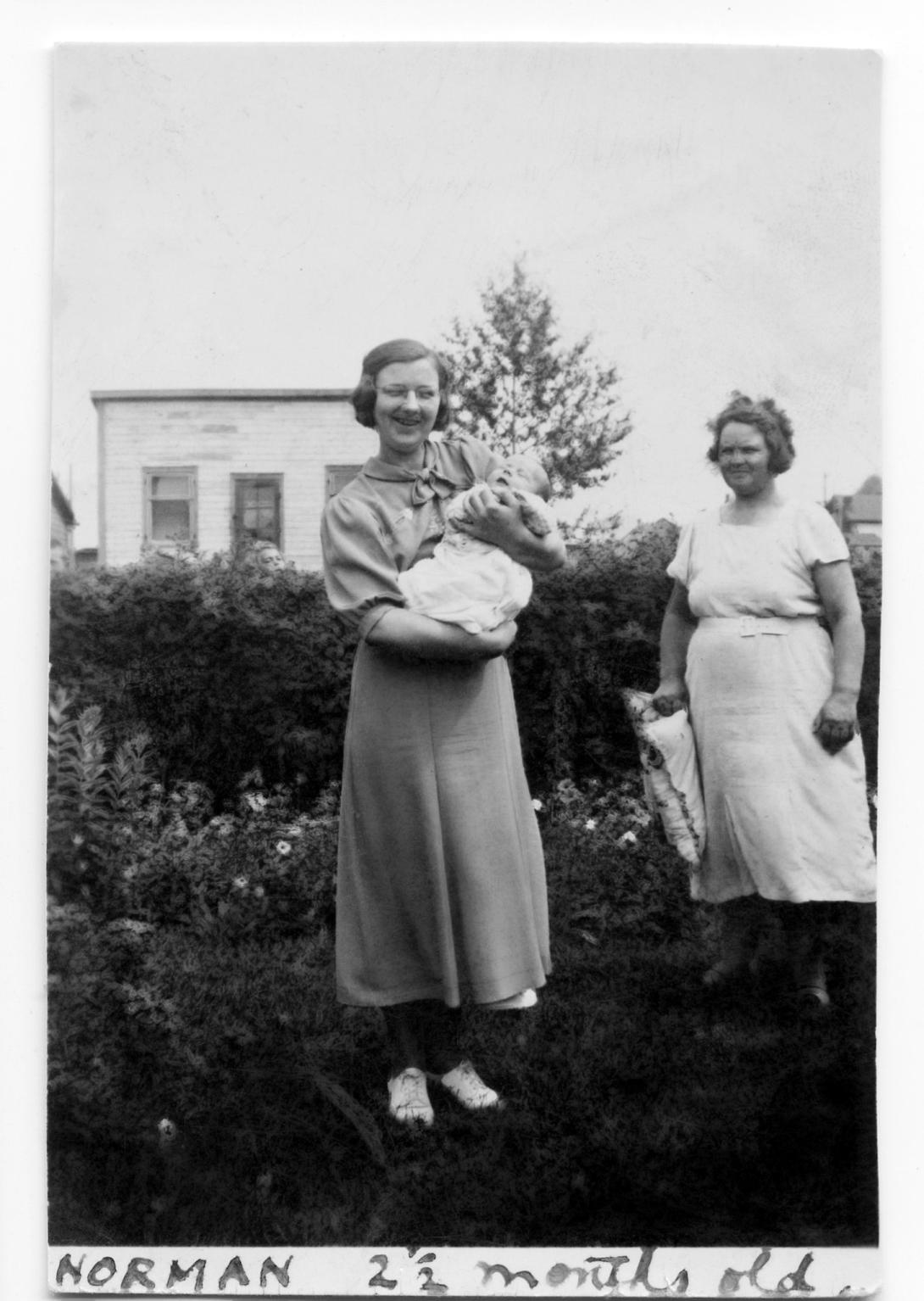 A black and white photo of a young woman holding and infant child. Beside her and slightly removed is an older woman. 