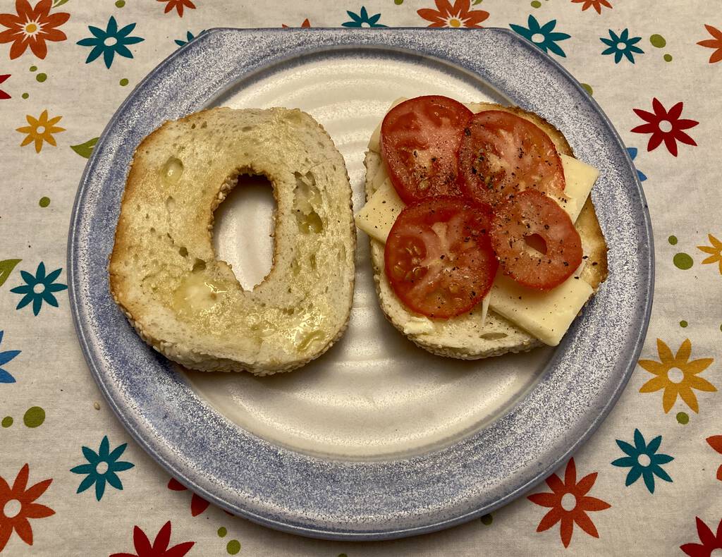 Photo of my lunch: cheese and tomato on a bagel, on a plate, set on a tablecloth.