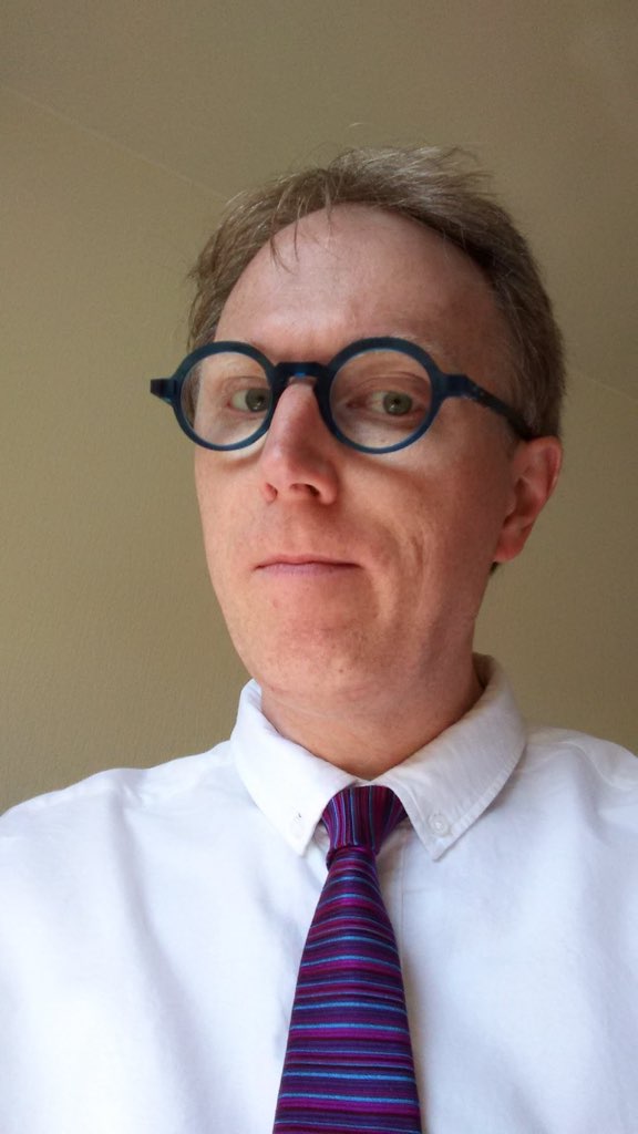 Me, wearing a white button-down shirt, purple tie, severe blue glasses.