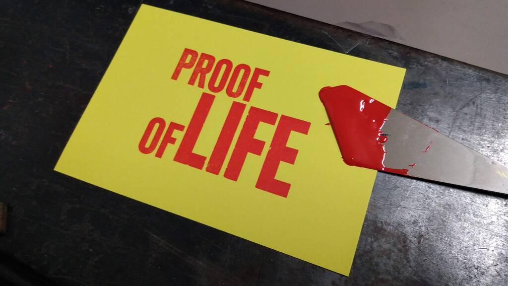 Proof of Life print, in red on yellow paper, with an ink spatula covered in red ink overlaid.