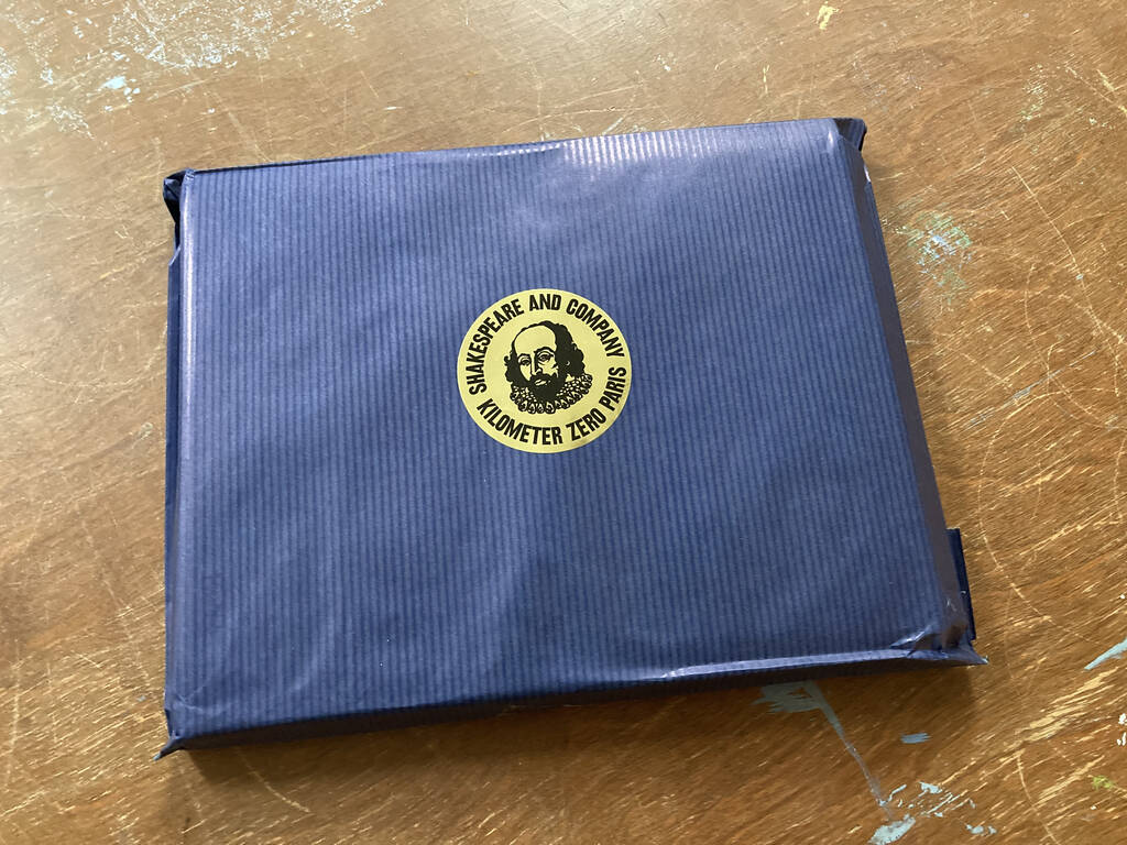 Book wrapping in a navy blue paper bag, with a round, gold Shakespeare and Company sticker on the front.