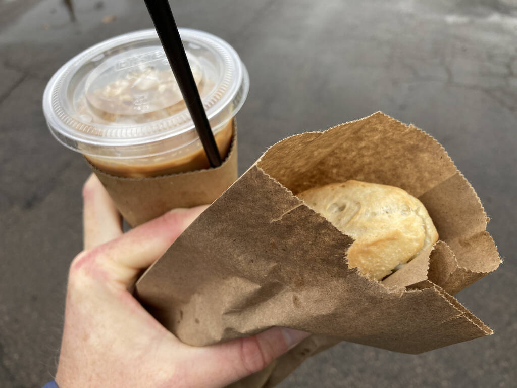 Photo of my left hand holding a plastic cup, with cover and straw, holding brown iced coffee, and showing the tip of a sandwich in a paper bag.