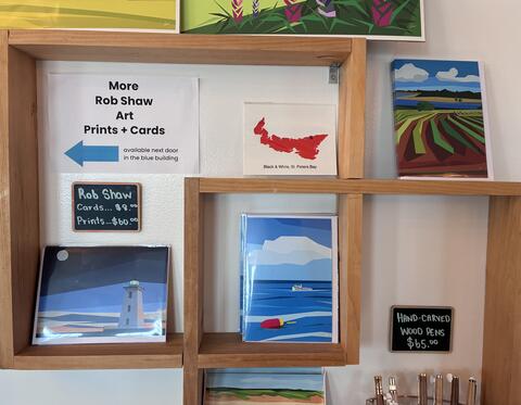 A shelf at Black & White, showing Rob Shaw postcards and notebooks (colourful, abstract) and one postcard, a red image of PEI with a black star showing Black & White.