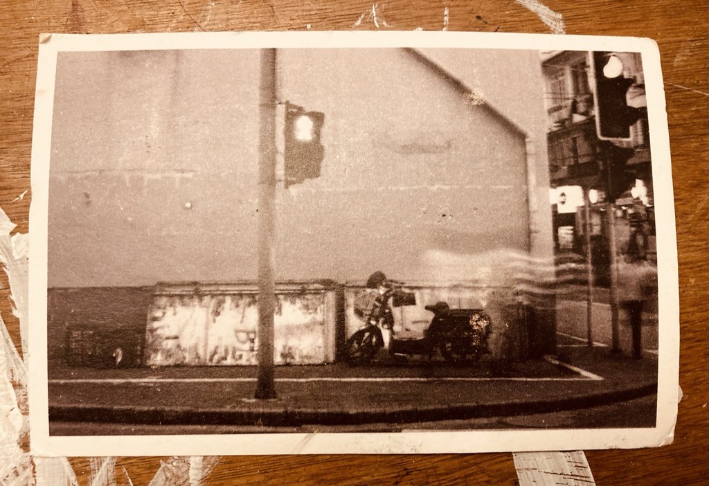 A blurry black and white postcard of a street corner in Shanghai at night, featuring a traffic light, a motor scooter, and bust street activity.